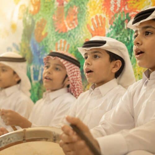 Four boys in traditional Qatari dress singing and playing hand drums in front of a colourful handprint mural