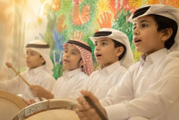 Four boys in traditional Qatari dress singing and playing hand drums in front of a colourful handprint mural