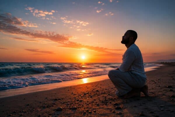 A man in white linen kneeling on the shoreline at sunset, looking out over the sea