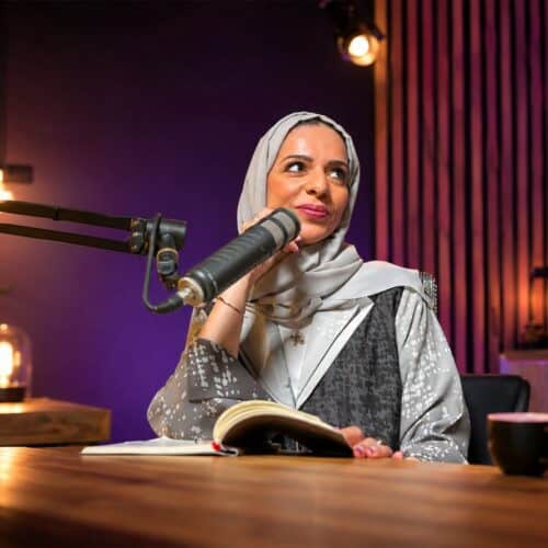 Safia Al Shehi reading from a script at a studio microphone, in a purple-lit recording booth
