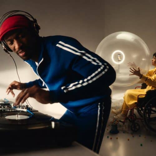 A DJ in a blue tracksuit and red headwrap mixing on turntables while a woman in a yellow abaya sits in a wheelchair beside a large illuminated sphere