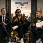 Three sonic branding agency archetypes waiting outside a pitch room: a corporate executive with binders of data, a starstruck music-industry connector in a leopard-print jacket, and an independent creative with headphones and a notebook of ideas