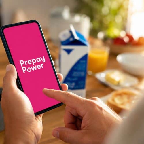 A hand holding a phone showing the Prepay Power app on a pink screen, at a sunlit breakfast table